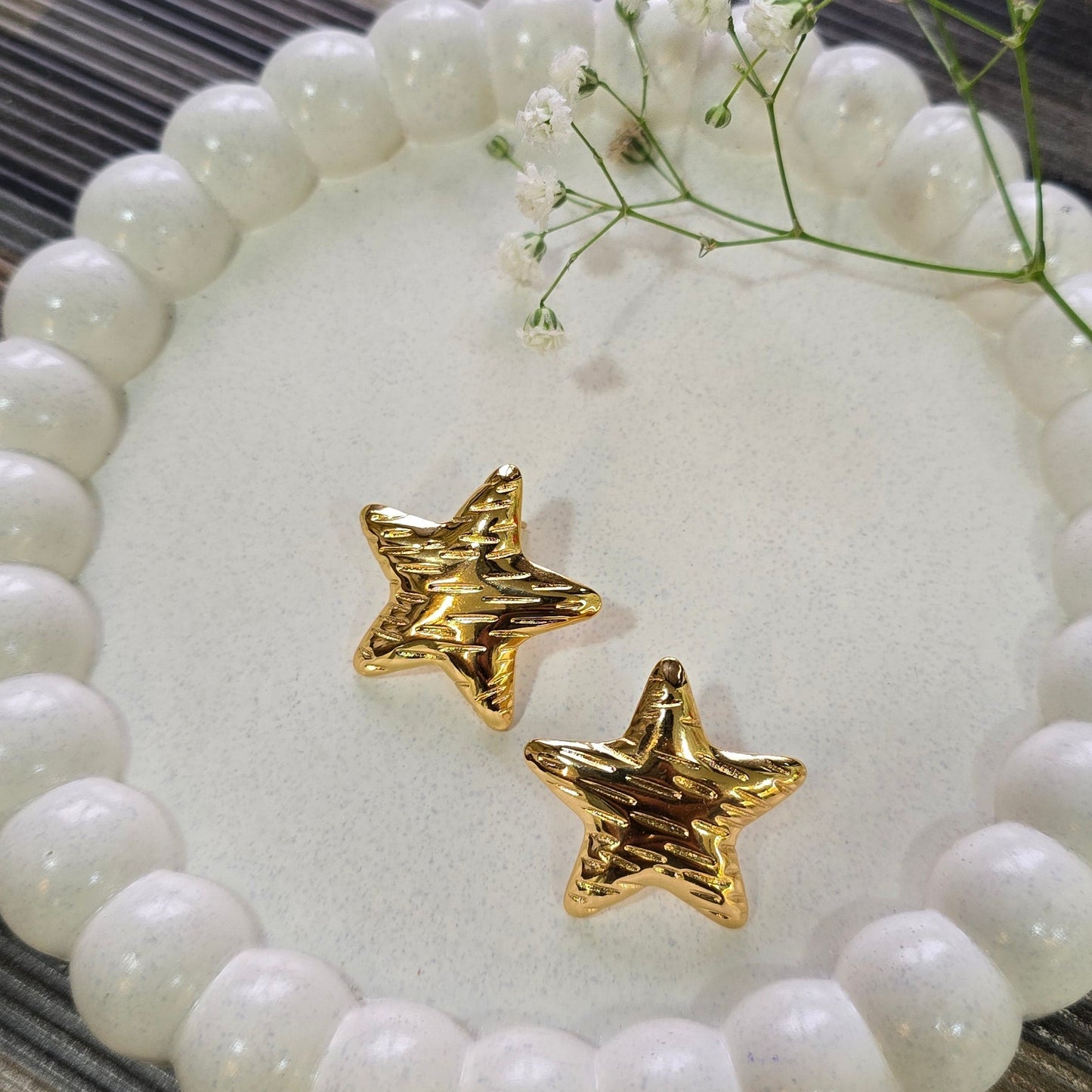 Gold star-shaped earrings on a white beaded dish with flowers on a wooden surface